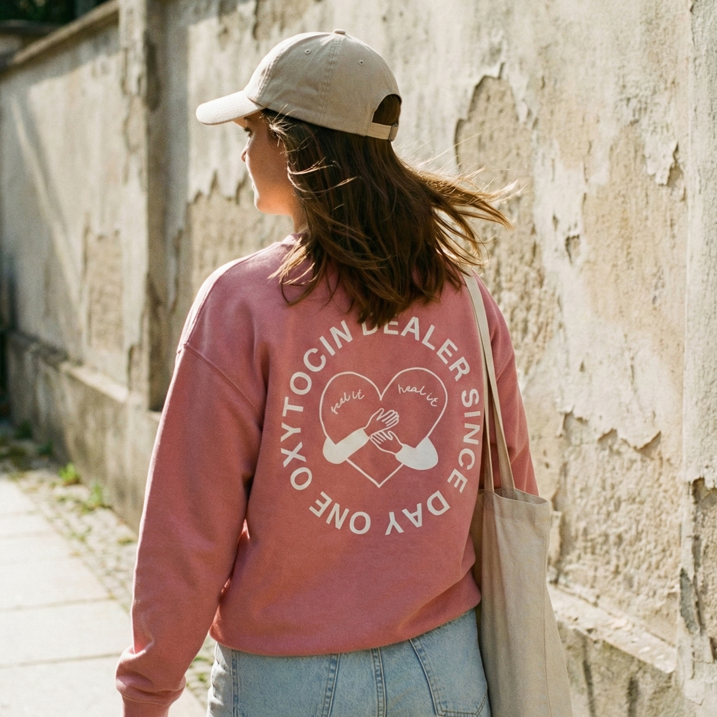 Frau mit Cap blickt nach vorne. Rückenaufnahme vom Sweatshirt in Pink und dem Design "Oxytocin Dealer Since Day One" von KOMM KLAR DIGGA